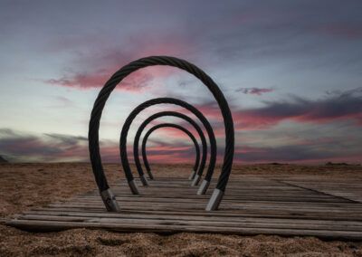 playa, pasarela, madera, aros, círculos, atardecer, cielo rosa, nubes, arena, costa, mar, horizonte, perspectiva, simetría, composición, paisaje marino, luz suave, crepúsculo, fotografía artística, profundidad, estructura, exterior, calma, colores pastel, minimalismo.