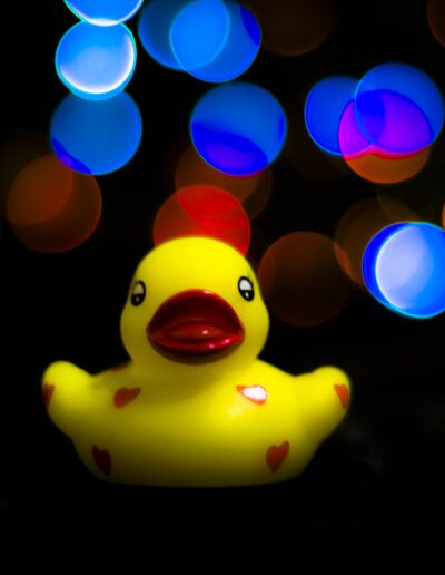 patito de goma, pato amarillo, juguete, infancia, diversión, colorido, luces desenfocadas, bokeh, fantasía, creatividad, alegría, inocencia, colores vivos, azul, rojo, violeta, amarillo, fotografía artística, composición creativa, juguete infantil, humor, entretenimiento, minimalismo, fondo oscuro, contraste, luces nocturnas, imaginación, felicidad, arte visual, decoración