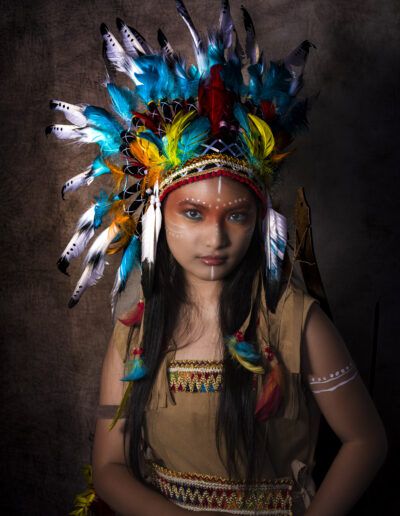 retrato, joven, tocado de plumas, maquillaje artístico, vestuario temático, colores, estudio, fondo oscuro, mirada, fantasía, personaje, iluminación dramática, plumas, cultura visual, fotografía artística.