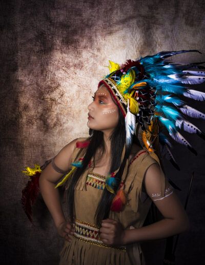retrato de perfil, joven, tocado, plumas, colores vivos, maquillaje facial, estudio fotográfico, fondo texturizado, pose, vestuario, fantasía, iluminación, fotografía artística, expresión, personaje.