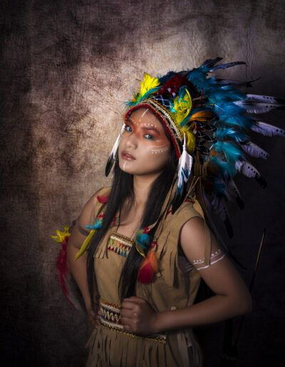 retrato femenino, joven, tocado de plumas, mirada, maquillaje artístico, colores, vestuario temático, estudio, fondo oscuro, expresión, fantasía, iluminación dramática, plumas, personaje, fotografía creativa.