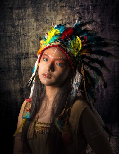 primer plano, joven, retrato, tocado de plumas, luz lateral, claroscuro, maquillaje, fondo oscuro, mirada intensa, estudio, fotografía artística, colores, vestuario, dramatismo, expresión.