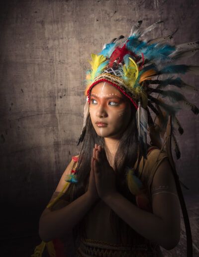 joven, oración, manos unidas, meditación, serenidad, tocado de plumas, retrato, maquillaje artístico, espiritualidad, contemplación, fondo oscuro, estudio, vestuario temático, paz, expresión.