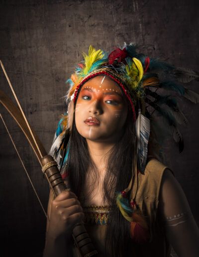 joven, arco, arquera, tocado de plumas, retrato, vestuario temático, personaje, aventura, fantasía, iluminación cinematográfica, estudio, maquillaje facial, fuerza, mirada, fotografía artística.