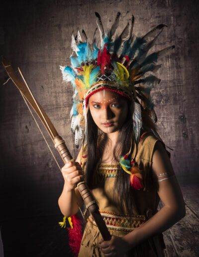 arquera, joven, arco de madera, tocado, plumas multicolor, retrato vertical, vestuario, fantasía, aventura, personaje, estudio fotográfico, iluminación dramática, maquillaje, determinación, fuerza.
