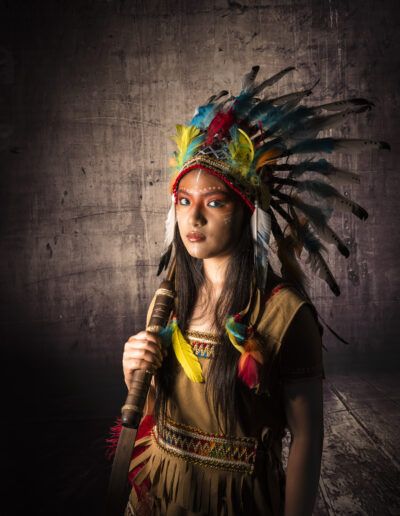 retrato, joven, arco, arquera, tocado de plumas, pose, estudio, fondo texturizado, vestuario temático, fantasía, aventura, maquillaje facial, iluminación, personaje, fotografía creativa.
