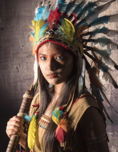 primer plano, joven, arquera, arco, tocado de plumas, retrato artístico, mirada intensa, luz cálida, claroscuro, maquillaje, estudio, personaje, vestuario, fantasía, expresión.