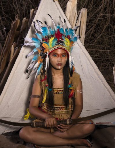 joven, arquera, arco, tocado de plumas, tienda, vestuario temático, retrato, pose sentada, maquillaje artístico, fantasía, colores, aventura, fotografía creativa, personaje, exterior.