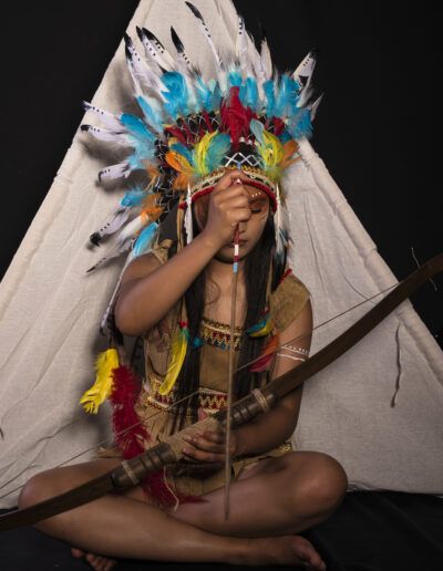 arquera, joven, arco y flecha, concentración, meditación, tocado de plumas, vestuario temático, pose sentada, maquillaje facial, retrato, estudio, fantasía, serenidad, personaje, fotografía artística.