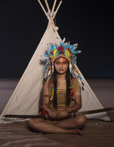 retrato frontal, joven, arco, tienda, tocado multicolor, plumas, simetría, fondo oscuro, vestuario temático, pose sentada, fantasía, personaje, maquillaje, estudio fotográfico, composición.