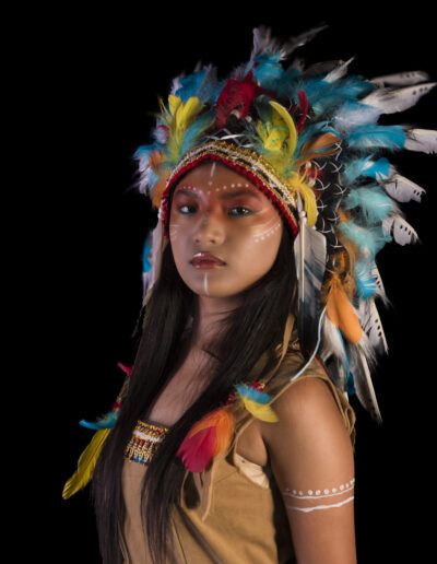 retrato, joven, tocado de plumas, fondo negro, maquillaje artístico, medio cuerpo, vestuario temático, colores vivos, mirada, estudio, iluminación, fantasía, personaje, fotografía creativa, expresión.