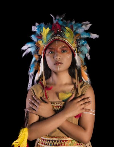 joven, brazos cruzados, gesto simbólico, tocado, plumas, retrato, fondo negro, maquillaje facial, vestuario temático, pose, fuerza, identidad visual, estudio, fotografía artística, expresión.