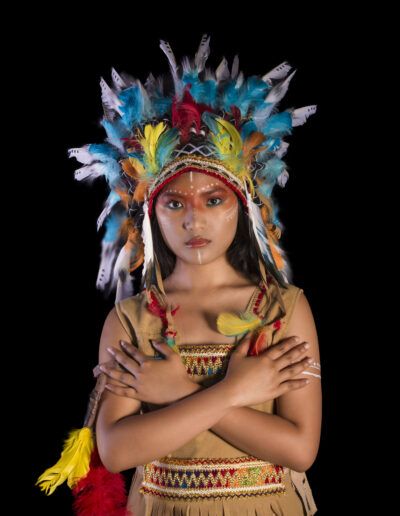 retrato frontal, joven, tocado multicolor, plumas, mirada directa, serenidad, fondo negro, maquillaje artístico, vestuario temático, estudio, colores vivos, simetría, personaje, expresión, fotografía de moda.