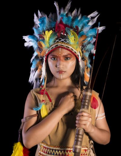joven, arquera, arco, flecha, tocado de plumas, mirada firme, retrato, fondo negro, vestuario temático, fuerza, aventura, personaje, maquillaje facial, estudio, fotografía artística.