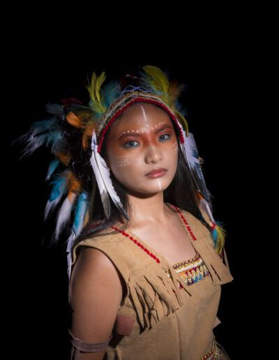 retrato tres cuartos, joven, tocado de plumas, maquillaje, fondo negro, iluminación suave, vestuario temático, mirada, pose, estudio fotográfico, fantasía, colores, elegancia, personaje, retrato artístico.