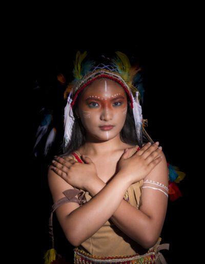 joven, brazos cruzados, retrato, tocado de plumas, mirada directa, gesto de protección, fortaleza, fondo negro, maquillaje artístico, vestuario temático, estudio, simbolismo, fotografía creativa, expresión, personaje.