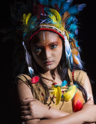 joven, retrato, brazos cruzados, tocado de plumas, fuerza, determinación, iluminación dramática, maquillaje artístico, fondo negro, vestuario temático, mirada intensa, carácter, personaje, estudio, colores.