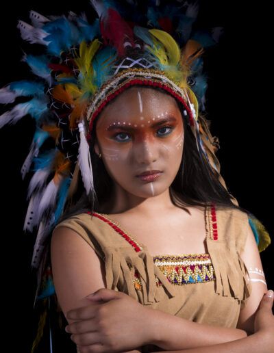 retrato frontal, joven, mirada desafiante, tocado, plumas, maquillaje facial, luz contrastada, fondo negro, expresión, fuerza, vestuario temático, estudio, fotografía artística, colores vivos, personaje.