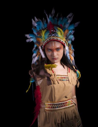 joven, mano extendida, gesto, movimiento, perspectiva, retrato, tocado de plumas, maquillaje artístico, fondo negro, pose teatral, energía, fantasía, personaje, expresión, fotografía creativa.