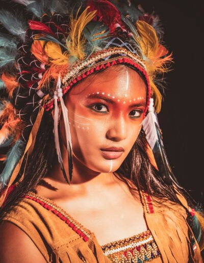primer plano, joven, tocado de plumas, luz cálida, mirada directa, maquillaje facial, retrato artístico, colores intensos, fondo oscuro, expresión, vestuario temático, iluminación, belleza, estudio, fotografía creativa.