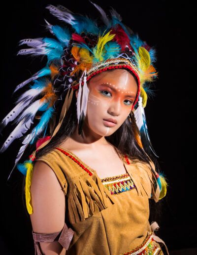 retrato, joven, medio cuerpo, tocado multicolor, plumas, pose elegante, fondo negro, maquillaje artístico, vestuario temático, mirada, colores vivos, estudio fotográfico, expresión, fotografía creativa, personaje.