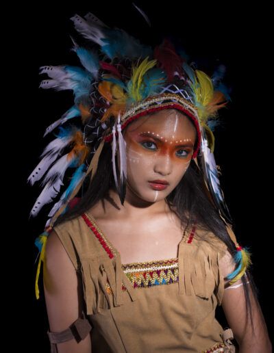 joven, retrato, tocado de plumas, serenidad, iluminación dramática, fondo negro, maquillaje facial, pose, vestuario temático, mirada, colores, estudio, expresión, fotografía artística, personaje.