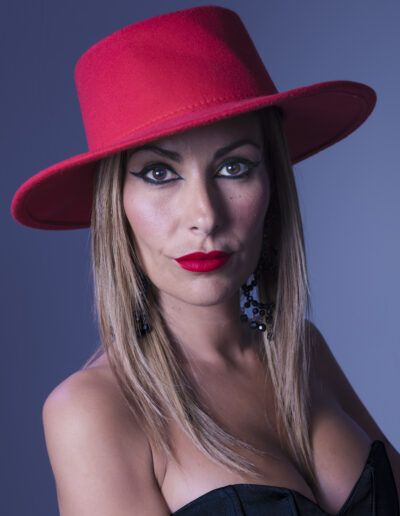 retrato frontal, mujer, sombrero rojo, ala ancha, labios rojos, maquillaje, fondo gris, moda, glamour, elegancia, mirada directa, pendientes, estudio, estilo, fotografía editorial.