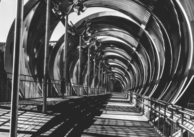 pasarela, arquitectura moderna, estructura metálica, blanco y negro, geometría, líneas, perspectiva, sombras, túnel, simetría, diseño urbano, profundidad, repetición, puente peatonal, abstracción arquitectónica.