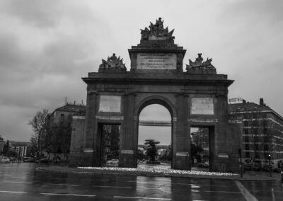 Puerta de Toledo, Madrid, monumento, lluvia, blanco y negro, arquitectura histórica, arco, calzada mojada, reflejos, glorieta, patrimonio, ciudad, cielo nublado, turismo, fotografía urbana.