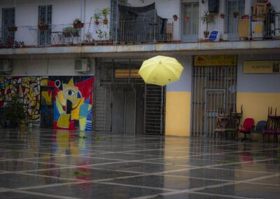 paraguas amarillo, lluvia, plaza, reflejos, suelo mojado, arte urbano, mural, escena callejera, color, ciudad, día lluvioso, contraste, arquitectura popular, vida cotidiana, fotografía urbana.