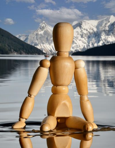 maniquí de madera, lago, montañas nevadas, reflejo, agua, paisaje, naturaleza, contemplación, tranquilidad, cielo azul, nieve, figura articulada, aventura, viaje, fotografía conceptual.