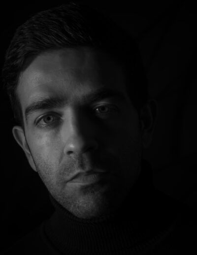 hombre, retrato masculino, blanco y negro, claroscuro, medio rostro, sombra, mirada frontal, primer plano, iluminación lateral, misterio, dramatismo, expresión seria, fondo negro, introspección, fotografía artística.